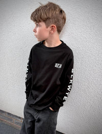 A young boy with light brown hair is shown in profile, wearing a black long-sleeve t-shirt with white text graphics on the chest and sleeves, paired with dark gray jeans, standing against a textured gray wall.