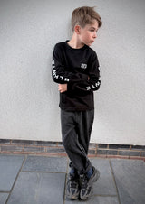 A young boy wearing a black long-sleeve graphic sweatshirt with white text on the sleeves, dark gray jeans, and athletic shoes, standing against a textured light gray wall.