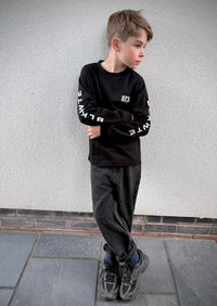 A young boy wearing a black long-sleeve graphic sweatshirt with white text on the sleeves, dark gray jeans, and athletic shoes, standing against a textured light gray wall.