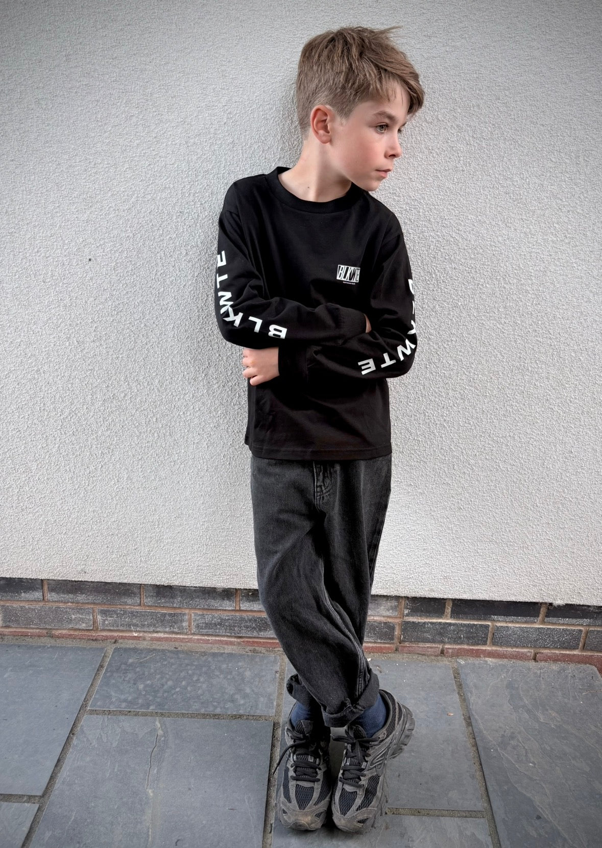 A young boy wearing a black long-sleeve graphic sweatshirt with white text on the sleeves, dark gray jeans, and athletic shoes, standing against a textured light gray wall.