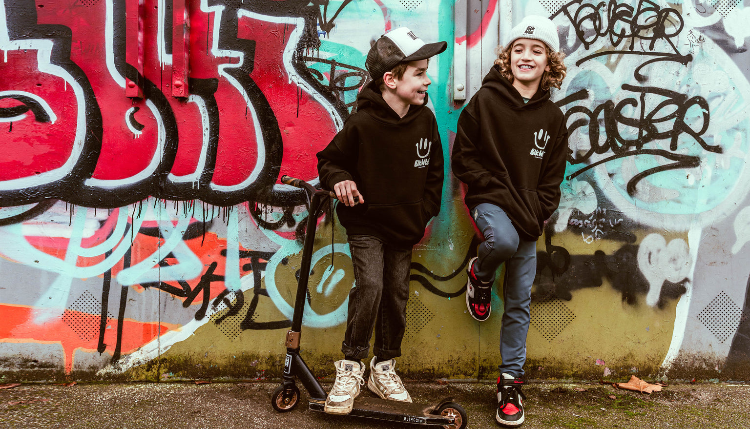 Two children with scooters in front of a graffiti-covered wall wearing BLKWTE hoodies