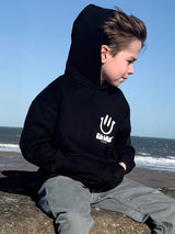 Child wearing a black hoodie with a logo sitting on a beach.
