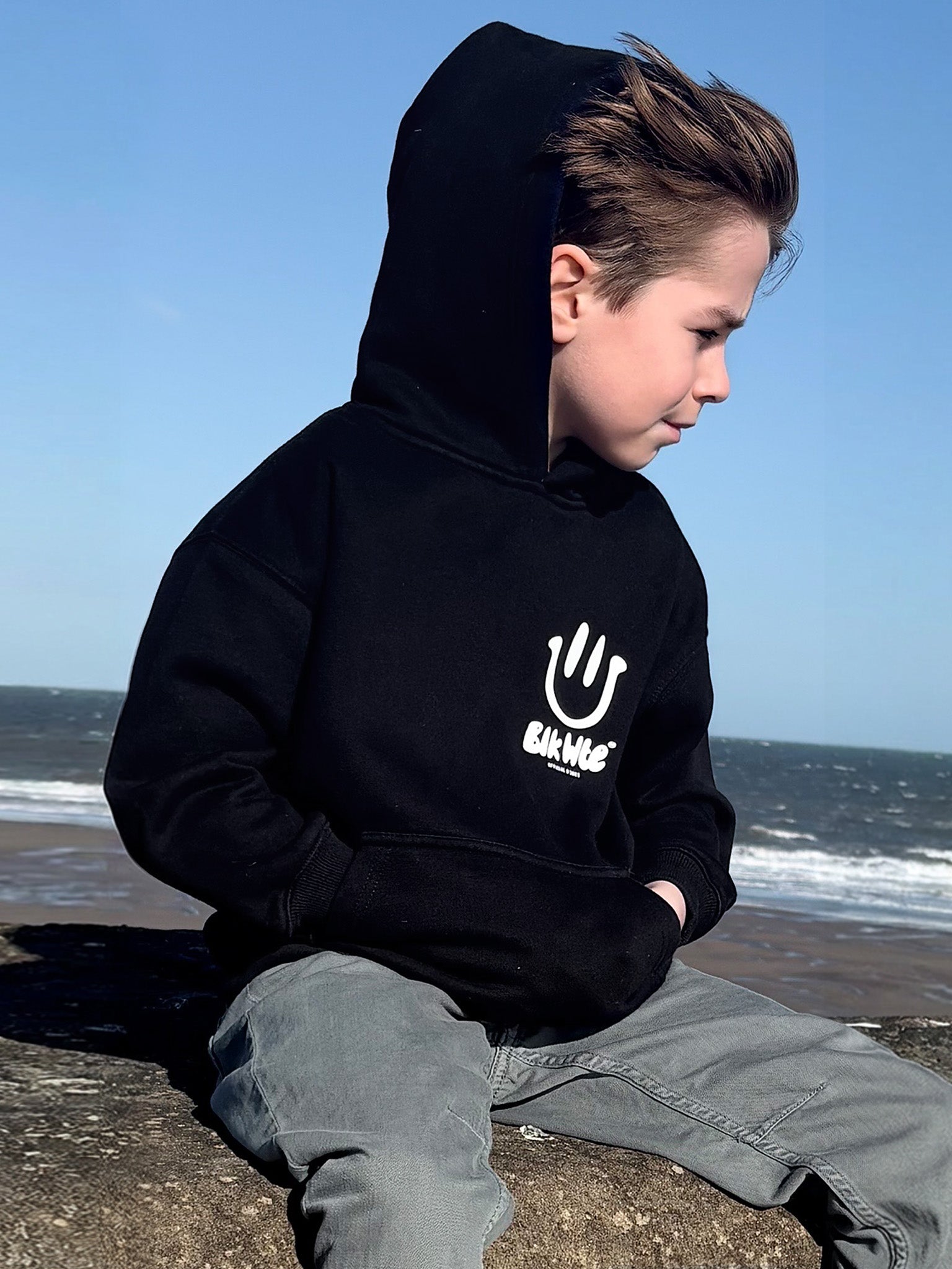 Child wearing a black hoodie with a logo sitting on a beach.